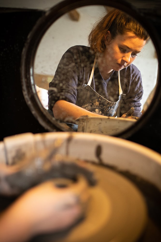 Manon Renault, céramiste près de Doué en Anjou et Saumur, en train de confectionner ses pièces en céramique artisanale sur un tour, avec son reflet dans le miroir au second plan, photographiée dans son atelier par Margot Vincent alias Margotine Photographies, photographe près d'Angers dans le maine-et-loire