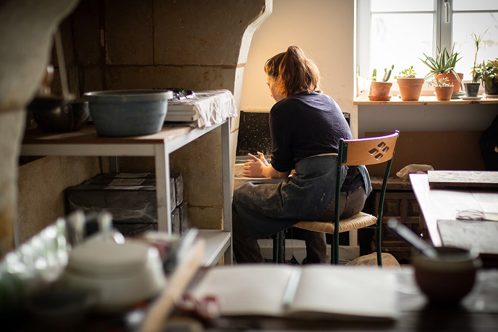 Manon Renault, céramiste près de Doué en Anjou et Saumur, vue de loin et de dos, en train de confectionner ses pièces en céramique artisanale sur un tour, photographiée dans son atelier par Margot Vincent alias Margotine Photographies, photographe près d'Angers dans le maine-et-loire